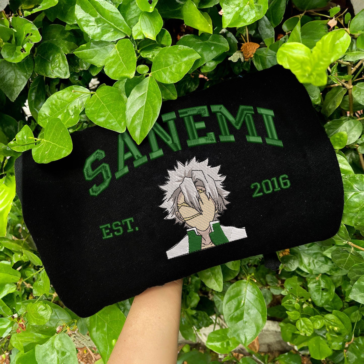 Black Sanemi embroidered sweatshirt held among green leaves.