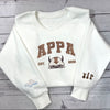 Folded white crewneck, gold “APPA 2005” embroidery