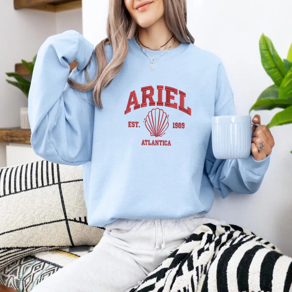 Woman Wearing Light Blue Ariel Embroidered Sweatshirt Holding Coffee Mug