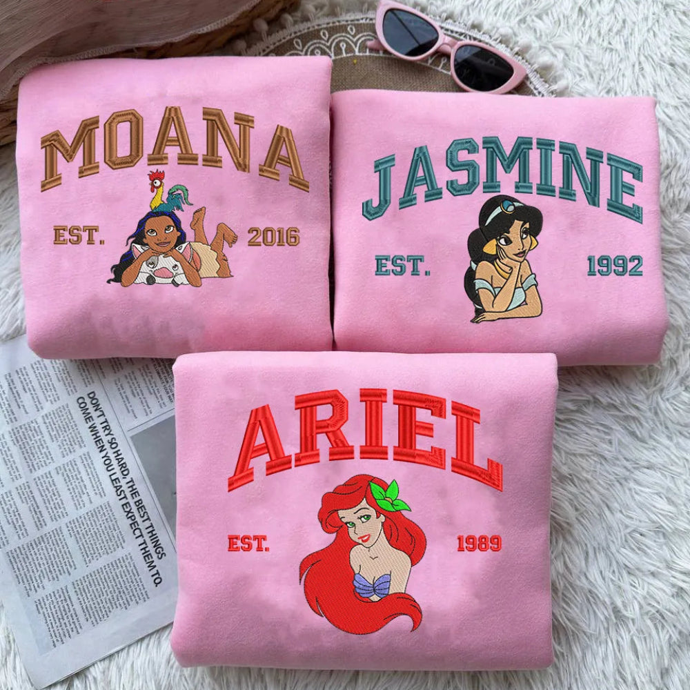 Folded Pink Sweatshirts Featuring Embroidered Moana, Jasmine, And Ariel Designs
